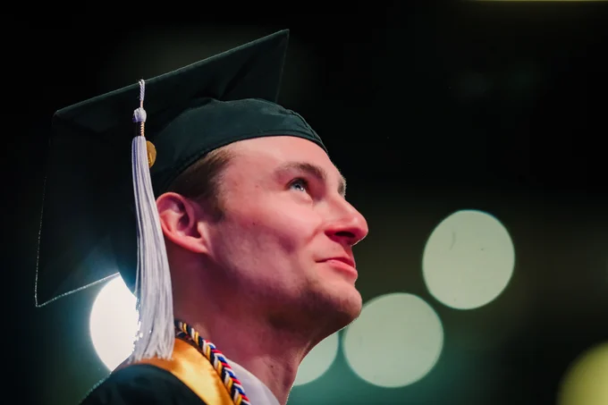 A close-up profile of a Kent State graduate looking upward with a quiet smile during the commencement ceremony, wearing a black cap with a white tassel and gold honor cord.