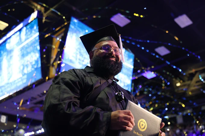 A Kent State graduate smiles and looks upward as gold and blue streamers rain down from the arena ceiling at the conclusion of the College of Arts and Sciences commencement ceremony, diploma in hand, surrounded by celebrating classmates and a packed arena.