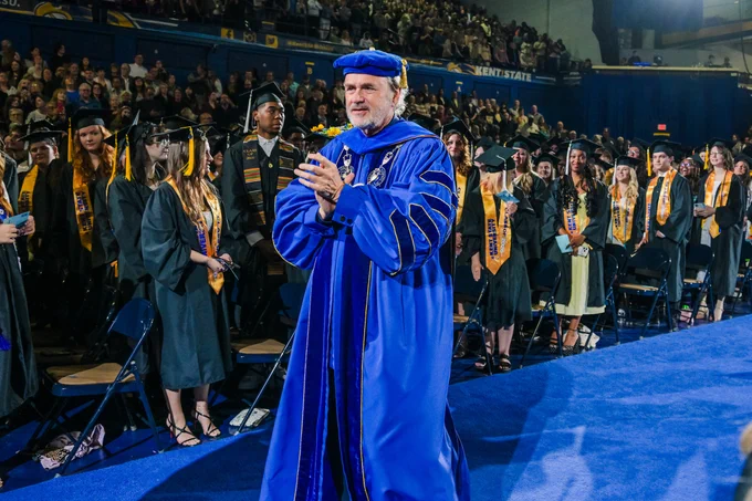Kent State University President Todd Diacon applauds while standing on the commencement stage in full blue presidential regalia, surrounded by graduates in black caps and gowns wearing gold Kent State stoles.