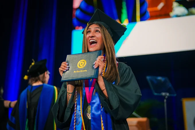 A Kent State graduate laughs with excitement while holding her diploma to her chest on stage at commencement, wearing black cap and gown with a blue honor stole and medal.
