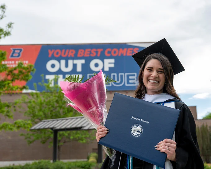 Boise State graduate student smiling outdoors with flowers and their diploma. 
