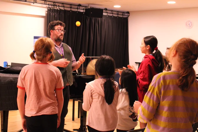 A music instructor demonstrates rhythm by tossing a ball to a group of attentive children in a classroom with a piano.