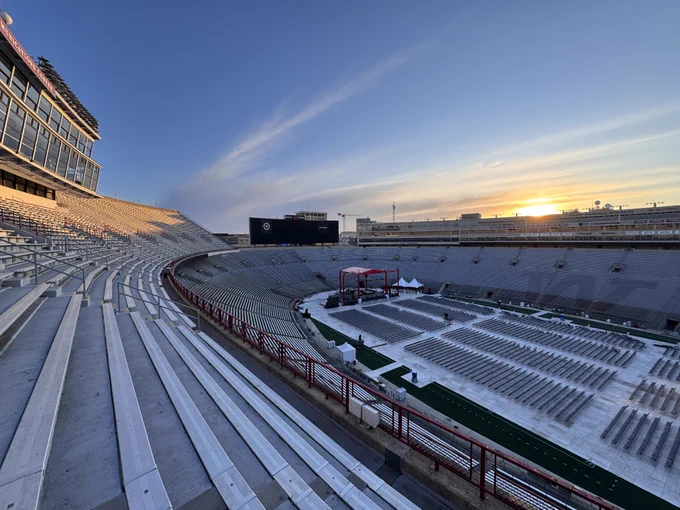 A wide shot of Camp Randall Stadium at sunrise, showing rows of empty bleachers and a stage setup on the field with floor seating for Spring 2026 Commencement.
