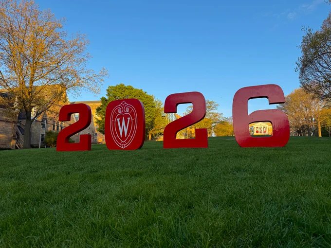 Large, red block numbers "2026" stand at the base of Bascom Hill at the University of Wisconsin–Madison, with the university's crest inside the zero and Bascom Hall visible through the opening of the six.