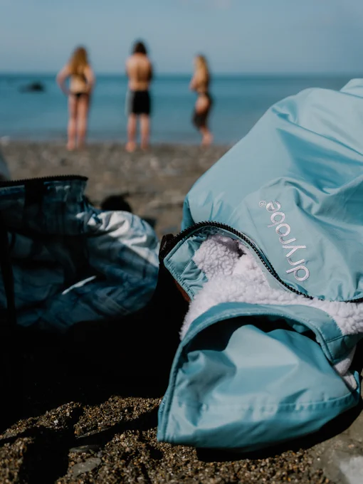 A close-up shot of a light blue Dryrobe® Advance change robe resting on a pebble beach. The focus is on the embroidered white Dryrobe® logo and the soft, white fleece lining. In the blurred background, three people stand near the ocean shore.