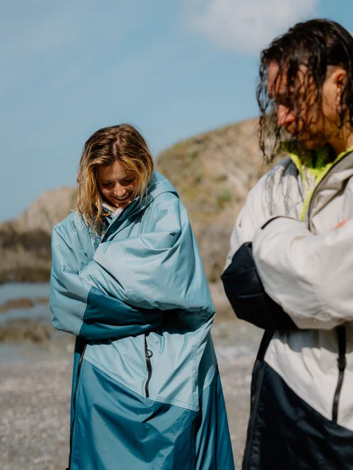 Two people on a pebble beach warming up after a swim. A woman in a two-tone blue Dryrobe® changing robe huddles into the oversized sleeves, smiling, while a man in a grey and black Dryrobe® stands beside her with his arms crossed.