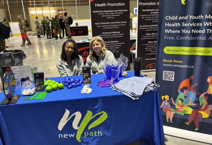 Two representatives sit behind a New Path health promotion table with informational materials and giveaways at a public event.