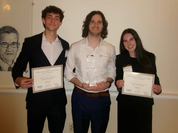 Three University of Florida students dressed in professional attire pose with certificates and a glass award after receiving the Best Paper Award at the 2026 IEEE Systems and Information Engineering Design Symposium.