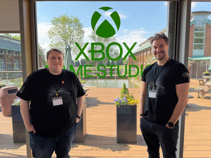 LokiJimmyB and Leator stand indoors in front of a large glass wall displaying the bright green Xbox logo and the words “Xbox Game Studios.”  Behind the glass, an outdoor deck with wooden flooring, planters filled with flowers and greenery, and a reflective water feature is visible. Buildings in the distance complete the backdrop, giving the scene a professional yet welcoming studio atmosphere.