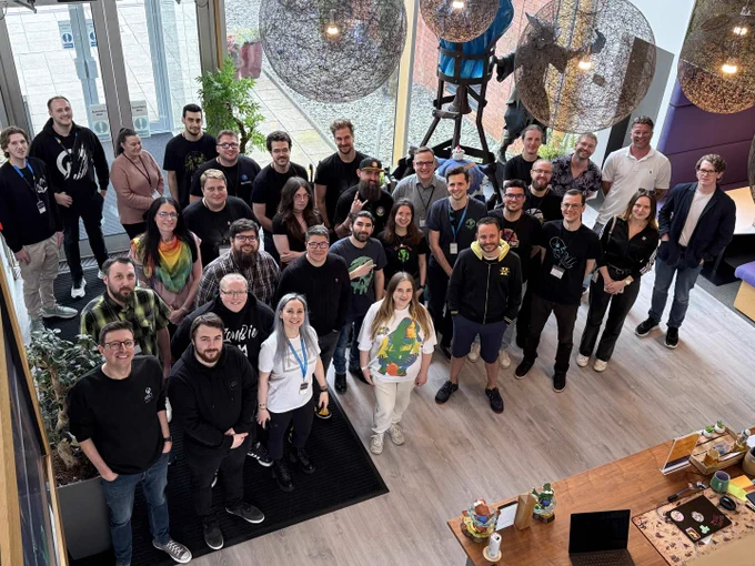 Large group photo taken inside a modern studio space, with around thirty people (developers, SoT Partners and AA leadership) standing and smiling toward the camera. The room has wooden floors, big windows, hanging spherical lights, plants, and a table with laptops and small figurines, giving the scene a creative, collaborative workspace atmosphere.
