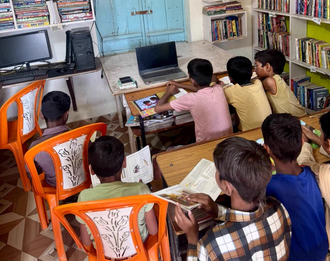 A group of children sitting together inside Bansa Community Library, quietly reading books and looking toward a laptop placed on a table. The room is filled with bookshelves, study materials, and colorful chairs, reflecting a shared learning and reading space in a rural community library.