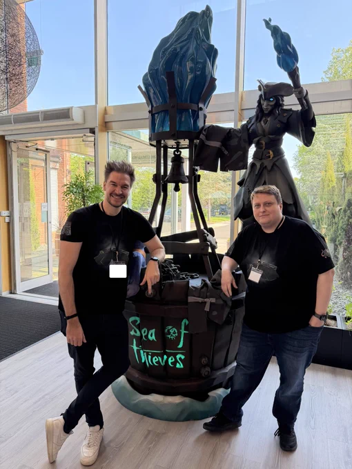 LokiJimmyB and Leator stand beside a large Sea of Thieves display featuring Belle, a glowing blue‑eyed pirate statue holding a torch in one hand and Obsidian Underpants over her other arm and a barrel-shaped structure with illuminated blue lettering. Bright natural light from tall windows fills the modern studio space behind them.