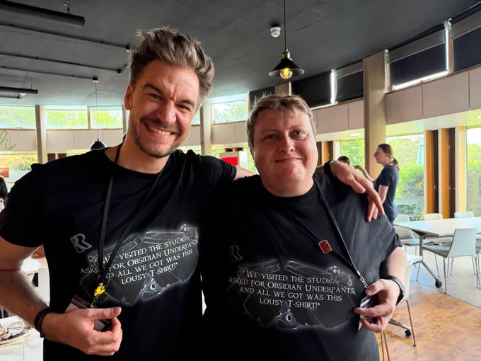 LokiJimmyB and Leator stand indoors wearing matching black T‑shirts which read: "We visited the studio, asked for Obsidian Underpants and all we got was this lousy T-Shirt". They’re smiling, with tables, chairs, and large windows in the softly lit background.