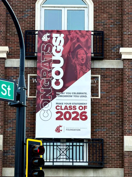 A large banner hangs on a brick building, featuring the logo of Washington State University. The text reads, "Congrats Cougs! Make your statement. Class of 2026. Today you celebrate, tomorrow you lead." A person is pictured cheering above the text.
