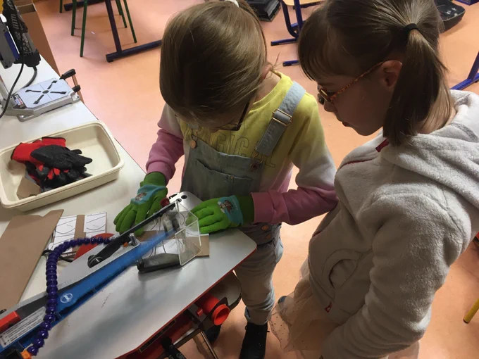 Deux élèves en classe utilisent une scie à chantourner pour découper une pièce de bois, l’une manipulant la machine avec des gants de protection pendant que l’autre observe attentivement à ses côtés.
