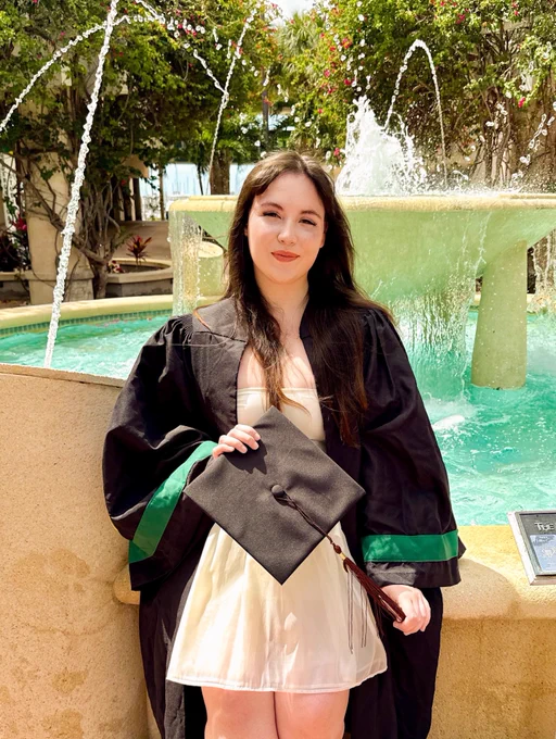 A person in a graduation gown holds a cap, standing in front of a fountain. The setting is sunny and surrounded by greenery and flowers.