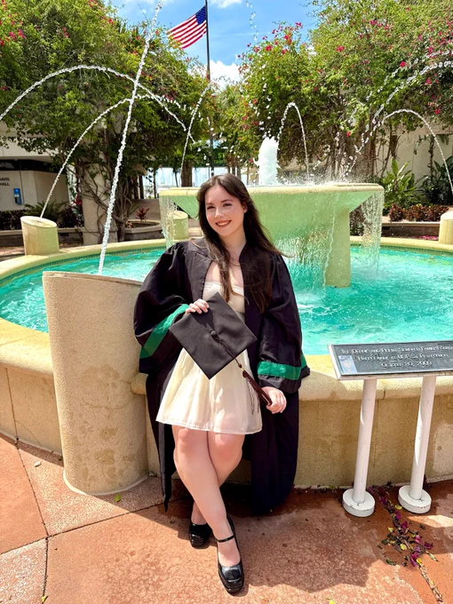 A person in a graduation gown sits on the edge of a fountain with water spraying upwards. They hold a graduation cap and wear formal attire underneath the gown. In the background, there are trees and an American flag.