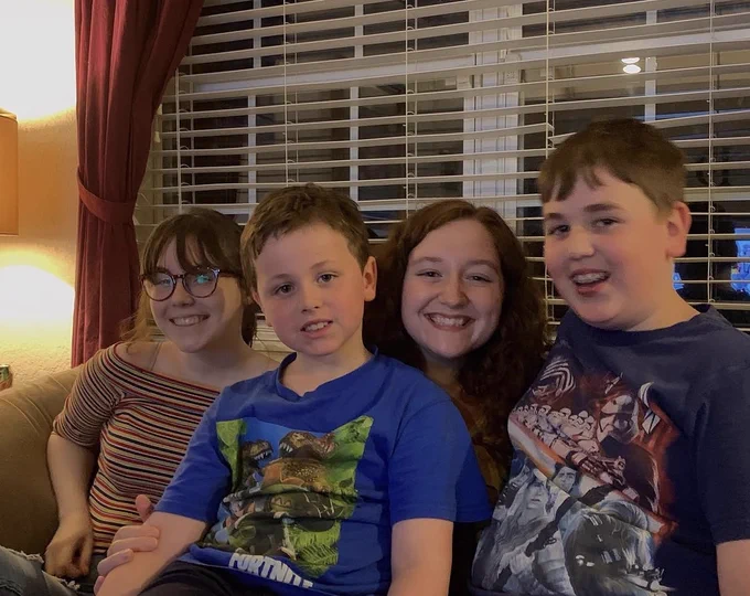 A group of four people sit close together on a couch. The older individuals are smiling broadly, while the younger ones look cheerful. One is wearing a striped shirt, and the other is wearing a brown top. The children are wearing shirts featuring characters from Fortnite and Star Wars. There is a window with blinds and curtains in the background.