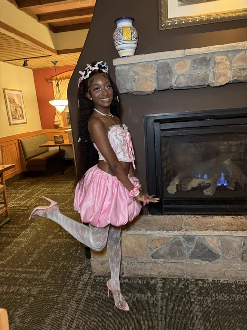 Pretty Black girl in pink and white corset top with bubblegum pink bubble skirt and lace stockings. Wearing pink stiletto heels. Pink, blue, and white bows in hair. Pink scrunchie and Apple Watch on wrist. Pretty Black girl is smiling and has one leg lifted up