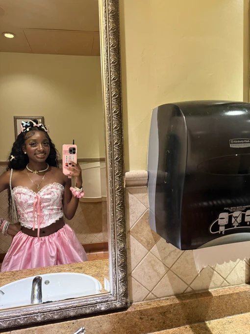 Pretty Black girl in pink and white corset top with bubblegum pink bubble skirt and lace stockings. Wearing pink stiletto heels. Pink, blue, and white bows in hair. Pink scrunchie and Apple Watch on wrist. Pretty Black girl is smiling in the mirror