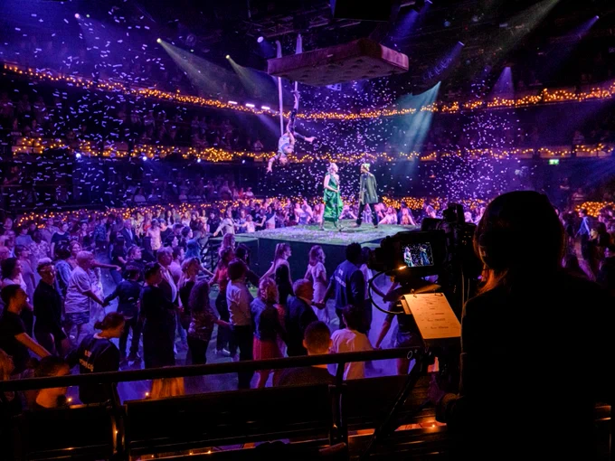 A group of people dancing on a stage surrounded by an audience. Lights surround the stage.
