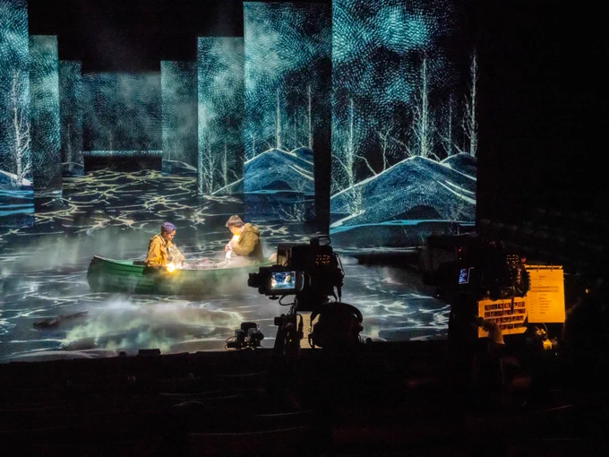 Two people in a boat on a stage with water patterns surrounding them. A camera focuses onto the stage.