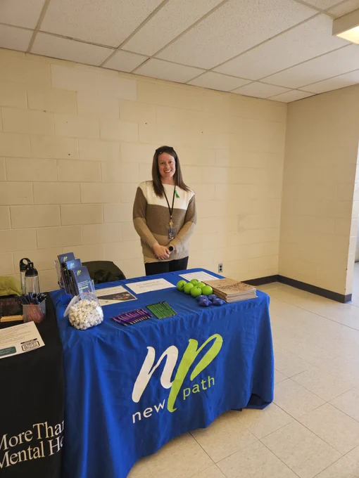 Woman standing behind a New Path informational table with brochures, green stress balls, and promotional items in a bright room.
