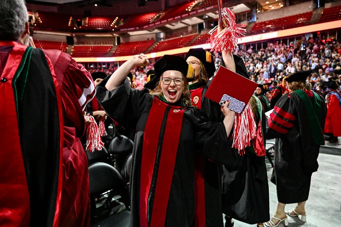 A joyful graduate in a cap and gown raises an arm in celebration while holding a diploma. The graduation ceremony is taking place in a large arena with an audience in the background.