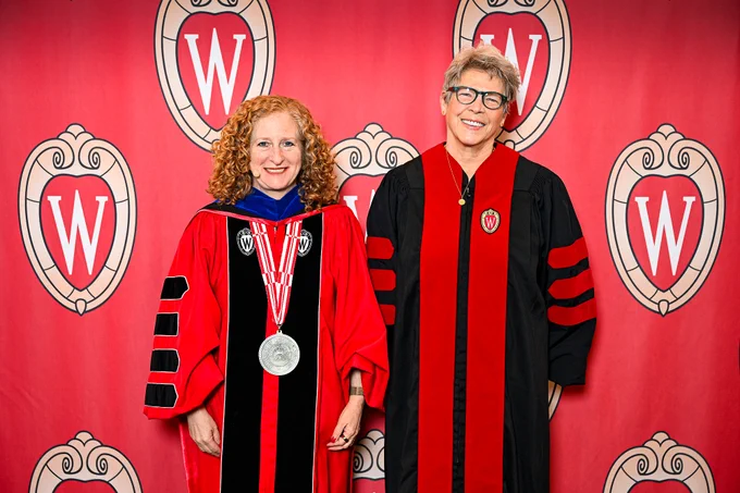 Chancellor Mnookin and honorary degree recipient Tia Nelson in academic robes shake hands in front of a backdrop featuring the University of Wisconsin-Madison logo.