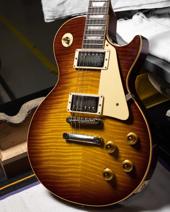 A Gibson Custom '59 Les Paul electric guitar with a sunburst finish, showcasing its iconic design and featuring dual humbucker pickups, a cream pickguard, and gold control knobs. It is resting on a table with some guitar accessories nearby.