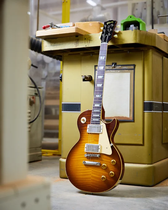 A Gibson Custom '59 Les Paul electric guitar with a sunburst finish is leaning against a piece of machinery in a workshop.