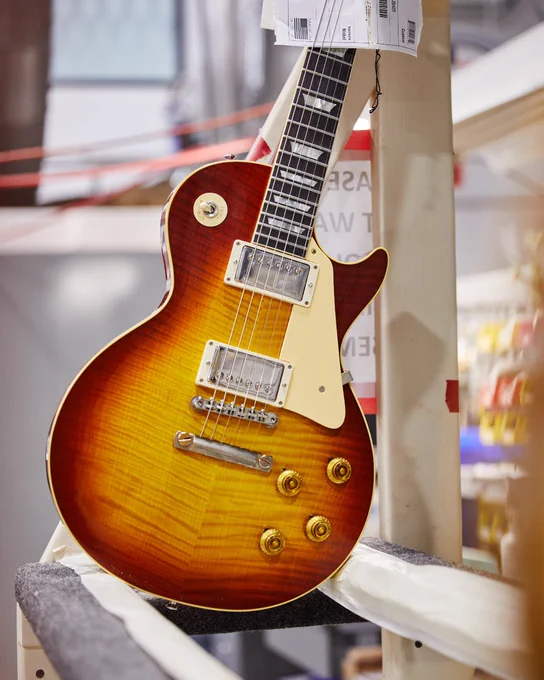 A sunburst Gibson Custom '59 Les Paul electric guitar, showcased vertically, hanging on a stand in a store or display setting.