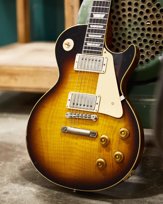 A Gibson Custom '59 Les Paul guitar with a sunburst finish leans against a wooden surface. The guitar features two humbucker pickups and four control knobs.