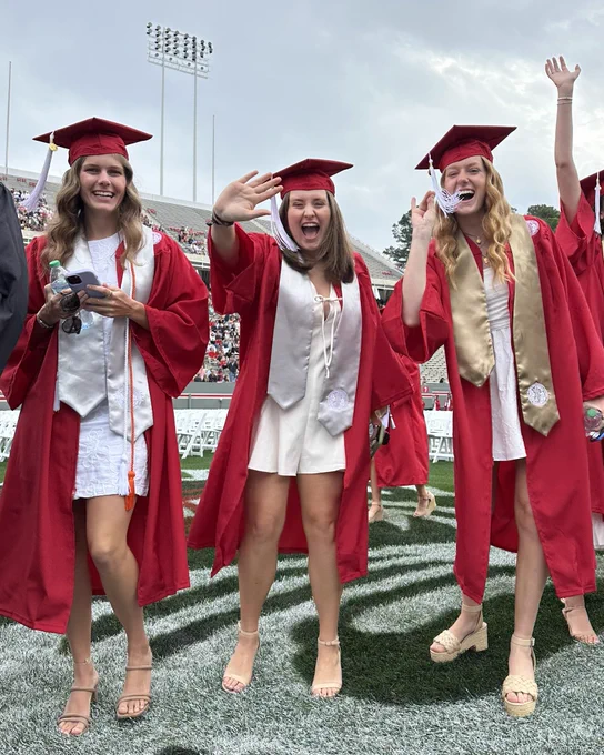 Three NC State graduates in red caps and gowns smiling and waving at the camera on the field in Carter-Finley Stadium.