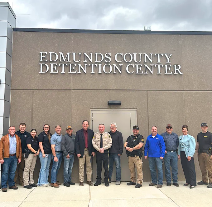 A group of thirteen people stands in front of the Edmunds County Detention Center building. They are dressed in a mix of casual and formal attire, with some in uniforms. The Edmunds County Detention Center sign is prominently displayed above them.