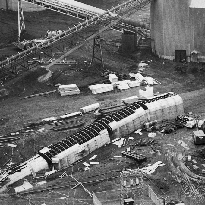 Black-and-white aerial view of the Westray mine disaster site, showing an industrial mine complex with large buildings, elevated conveyor belts, muddy ground, scattered construction materials, vehicles, equipment, and a long damaged cylindrical structure lying across the site.
