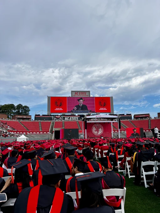 Graduates sit on the field at Carter-Finley Stadium while Harry Sideris speaks on stage.
