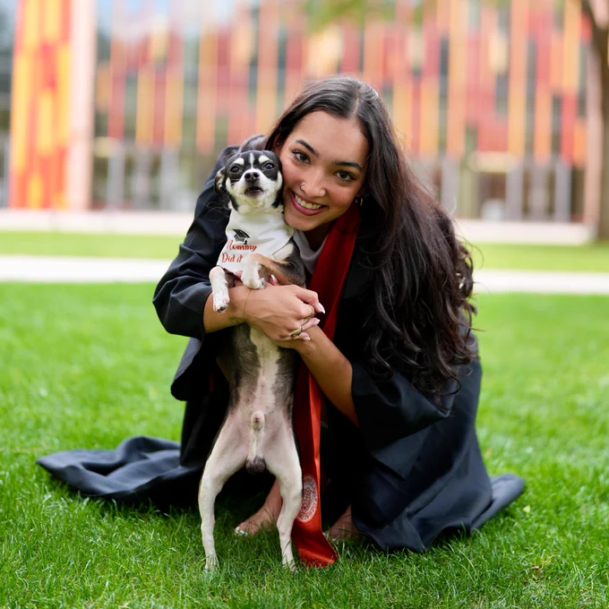 A person in a graduation gown is crouching on grass, smiling and holding a small dog wearing a bandana that reads "committed darling." A building with colorful vertical stripes is in the background.