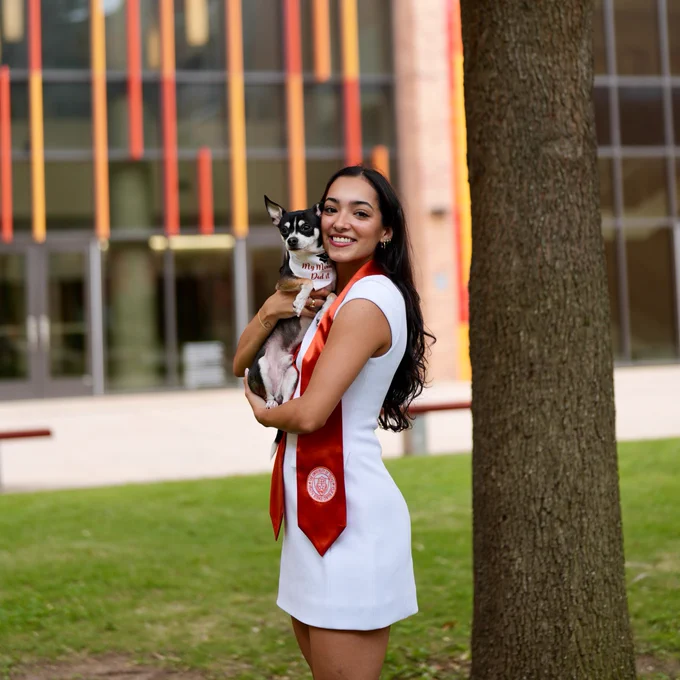 A person in a white dress and red stole is standing on grass, holding a small black and white dog wearing a "My Mom Did It" tag. The background features a building with vertical colorful panels.