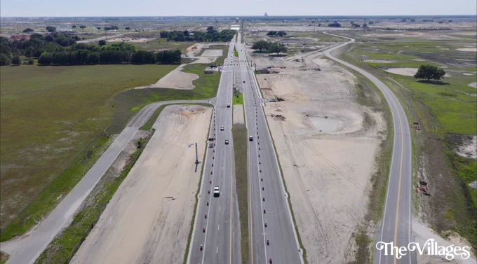 An aerial view showing a large highway under construction, surrounded by open fields and clear skies. The image captures a sense of expansiveness and infrastructure development.