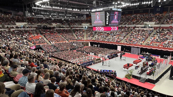 Color indoor photo showing Beasley Coliseum full of spectators in the stands at Spring commencement. A stage is in the front with a large video screen overhead. Hundreds of students sit in seats on the floor while wearing black, red or silver gowns with corresponding caps. 