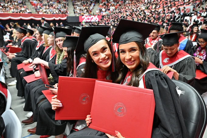Color indoor photo of 2 W S U grads holding diploma covers while looking at the camera. 
