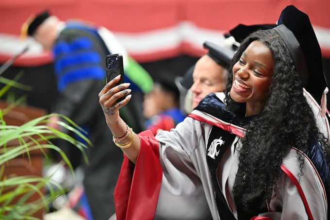 Color indoor photo of a grad in a gray gown while taking a selfie. 