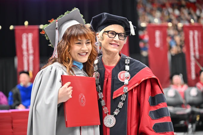 Color indoor photo of a student in a gray gown holding a crimson diploma cover standing next to W S U President Cantwell. 