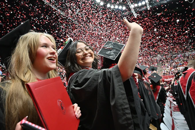 Color indoor photo of two grads taking a selfie with confetti falling from the ceiling. 