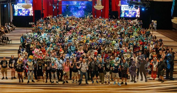 Photograph of the VRChat meetup taken at Furry Weekend Atlanta. Hundreds of furries, including fursuiters and plain-clothed people, are grouped together for a large group photo. The setting is a large, lavish ballroom with chandeliers hanging from the ceiling and television screns in the background.