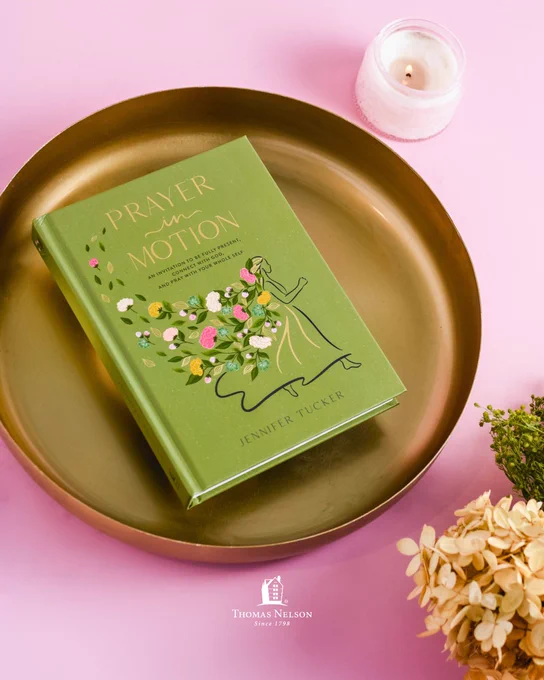 A green book titled "Prayer in Motion" by Jennifer Tucker rests on a round brass tray. A lit candle and some dried flowers are nearby. The background is a soft pink surface with the Thomas Nelson logo visible at the bottom.
