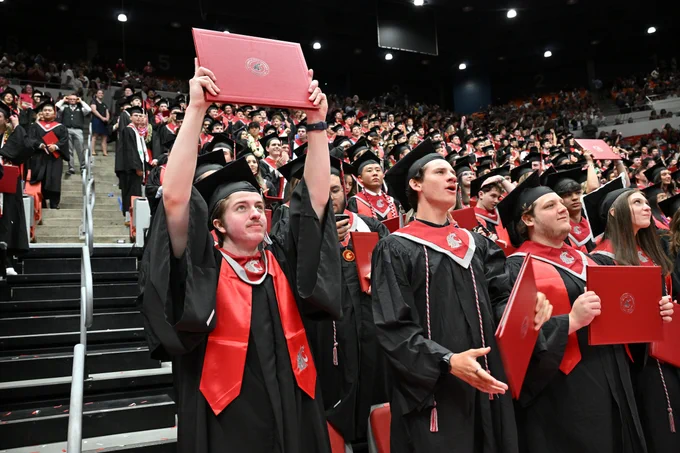 Color indoor photo of grads at W S U Pullman's graduation, with one holding a red diploma cover above their head. 