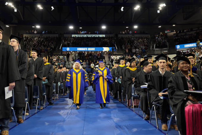 A graduation ceremony featuring individuals in academic regalia walking down a blue aisle toward the camera. The background shows an audience seated in a large indoor arena with congratulatory messages displayed on screens. The atmosphere is celebratory and formal.
