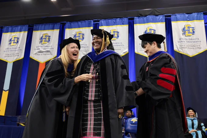 A person in academic robes is being hooded at a South Dakota State University ceremony. Two individuals, also in academic attire, are beside this person, who is smiling. Banners with the university's logo hang in the background.